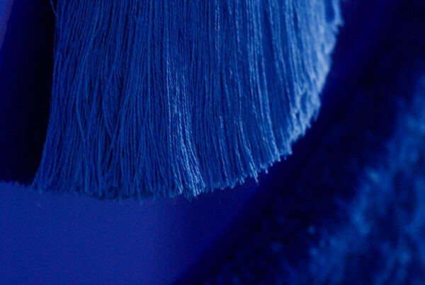 A close-up of an abstract, textured artwork in varying shades of blue. The piece features a long, flowing strand-like texture that contrasts against a deep blue background, creating a sense of movement and softness.
